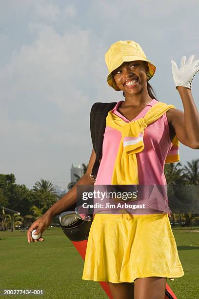 young woman carrying golf bag on golf course, waving and smiling - golfhandschuh stock-fotos und bilder