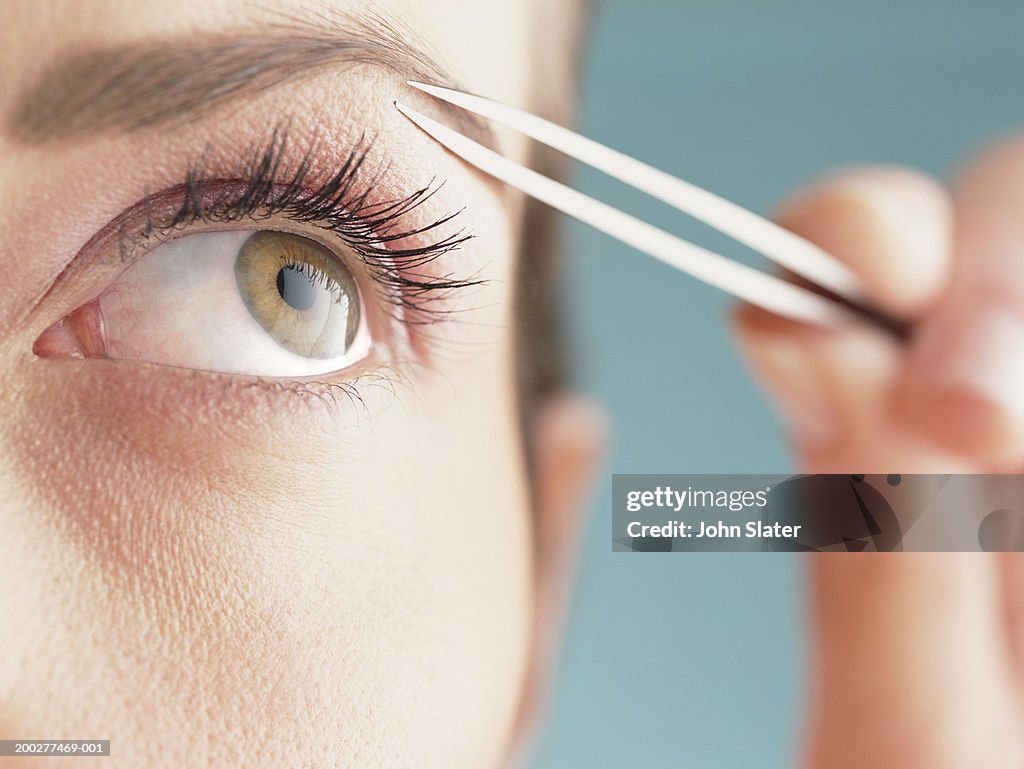 Young woman shaping eyebrow with tweezers, close-up of eye