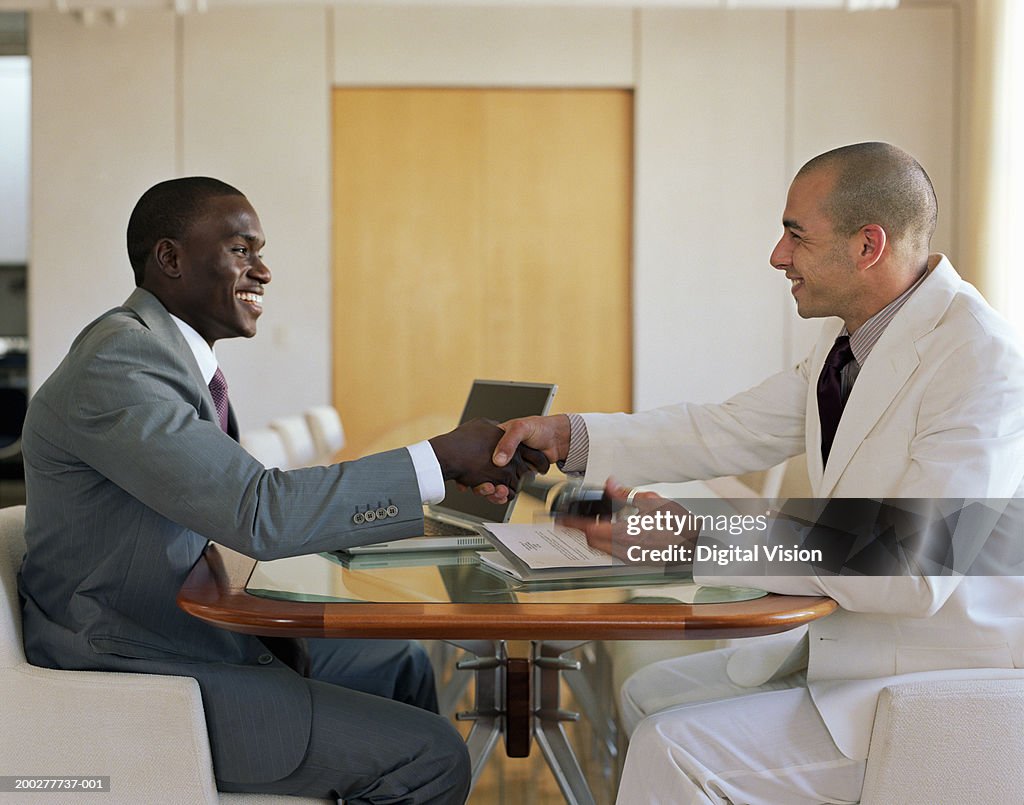 Two businessmen shaking hands across table, smiling, side view
