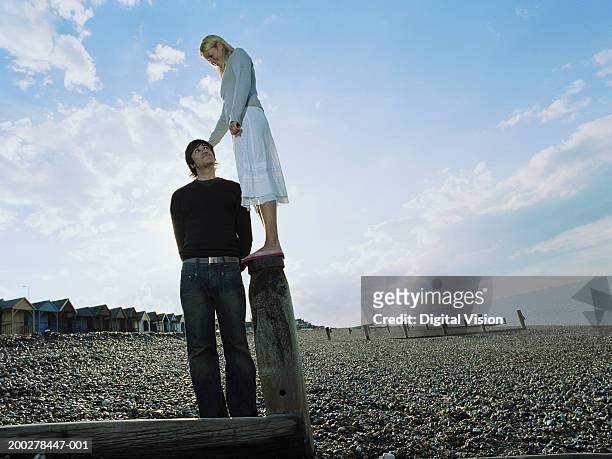 couple on pebbled beach, man looking up at woman standing on groyne - basso foto e immagini stock