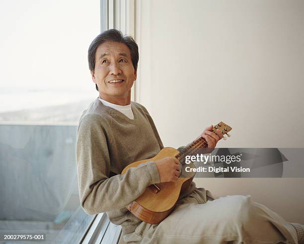 senior man sitting by window playing ukelele, looking up - japanese ol stockfoto's en -beelden