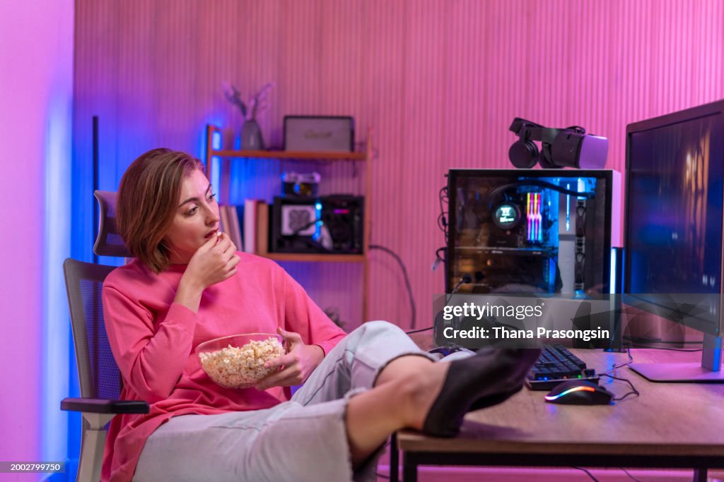 Smiling young blonde eating popcorn, while sitting in front of her computer