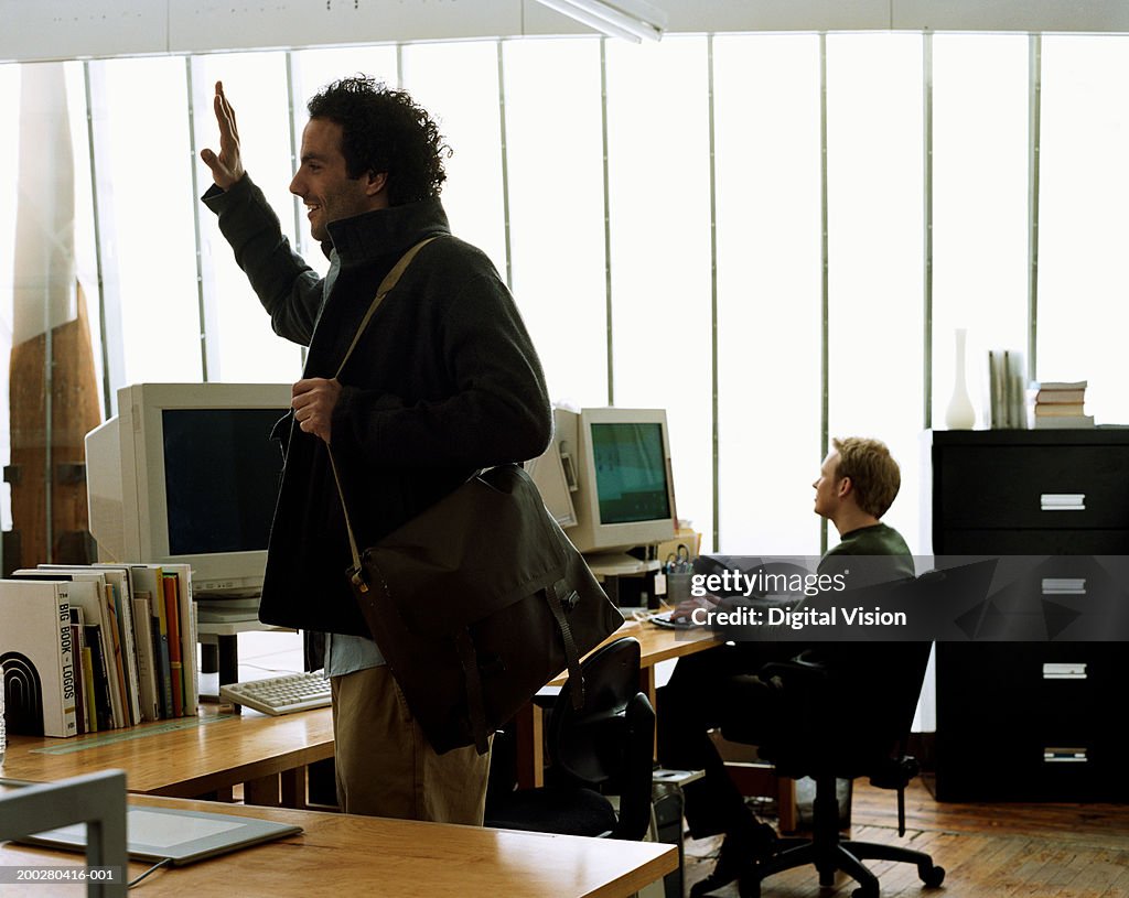 Man standing in office carrying shoulder bag, hand raised