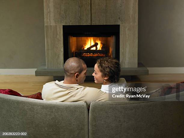 young man and woman embracing on sofa beside fireplace, rear view - sitting by fireplace stock pictures, royalty-free photos & images