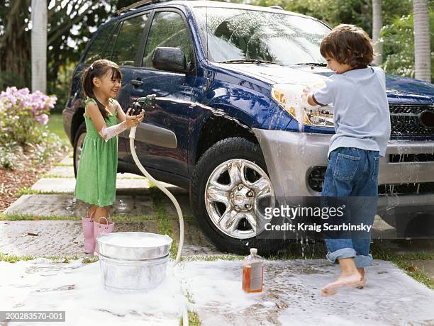 girl (4-6) aiming hose at boy (5-7) washing car on driveway, smiling - girls taking a showering stockfoto's en -beelden