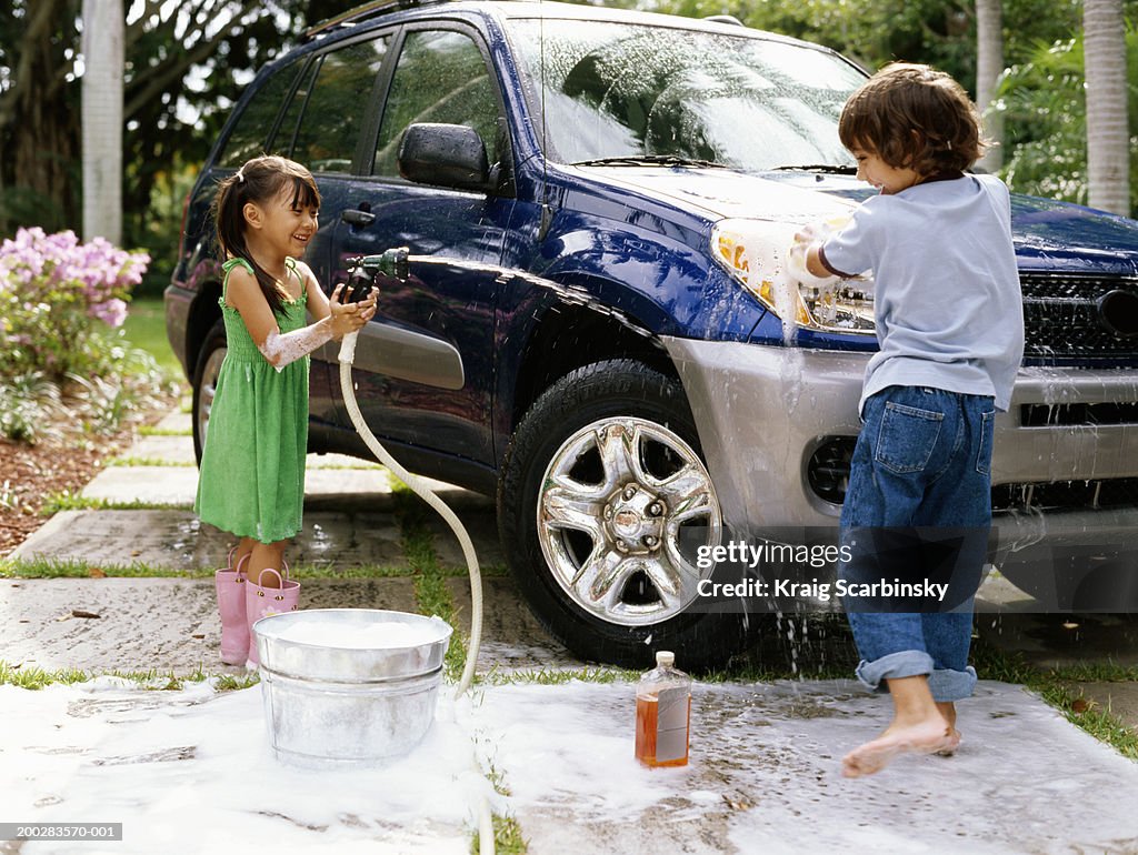 Girl (4-6) aiming hose at boy (5-7) washing car on driveway, smiling