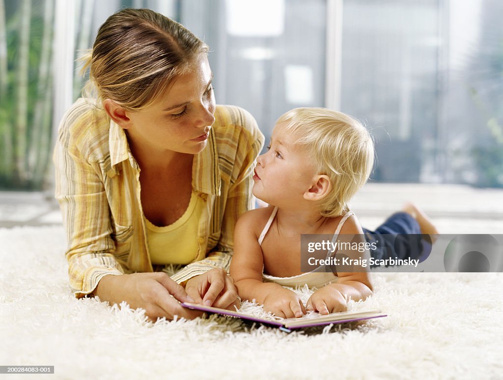 Woman and female toddler (21-24 months) lying on rug with book, looking at each other
