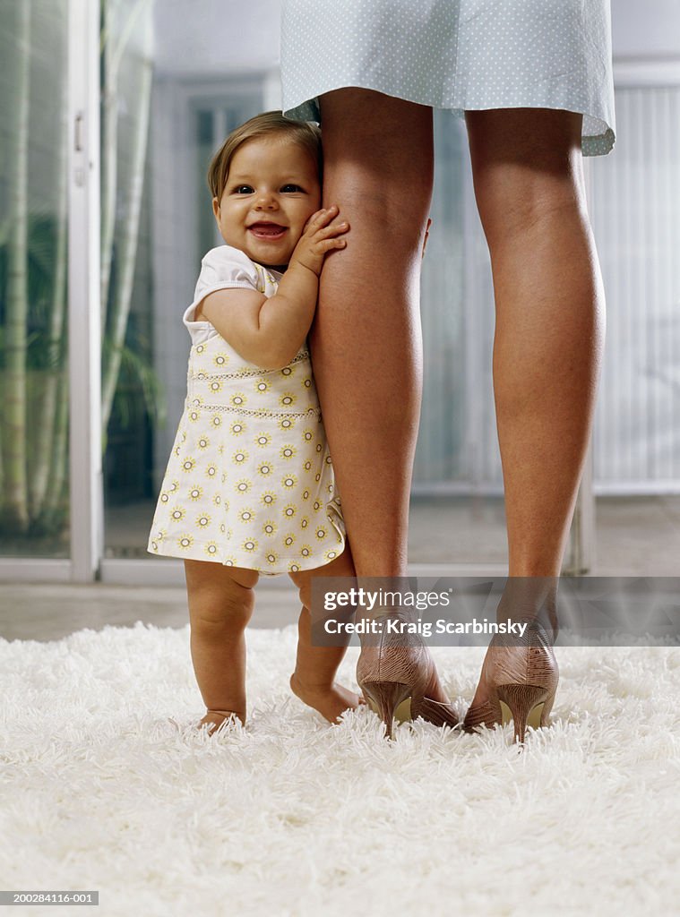 Baby girl (6-9 months) standing on rug, holding onto mother's legs