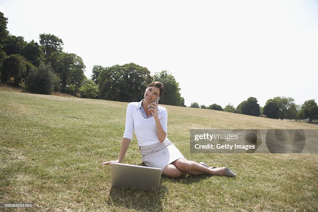 Businesswoman sitting on lawn by laptop, using mobile phone, smiling