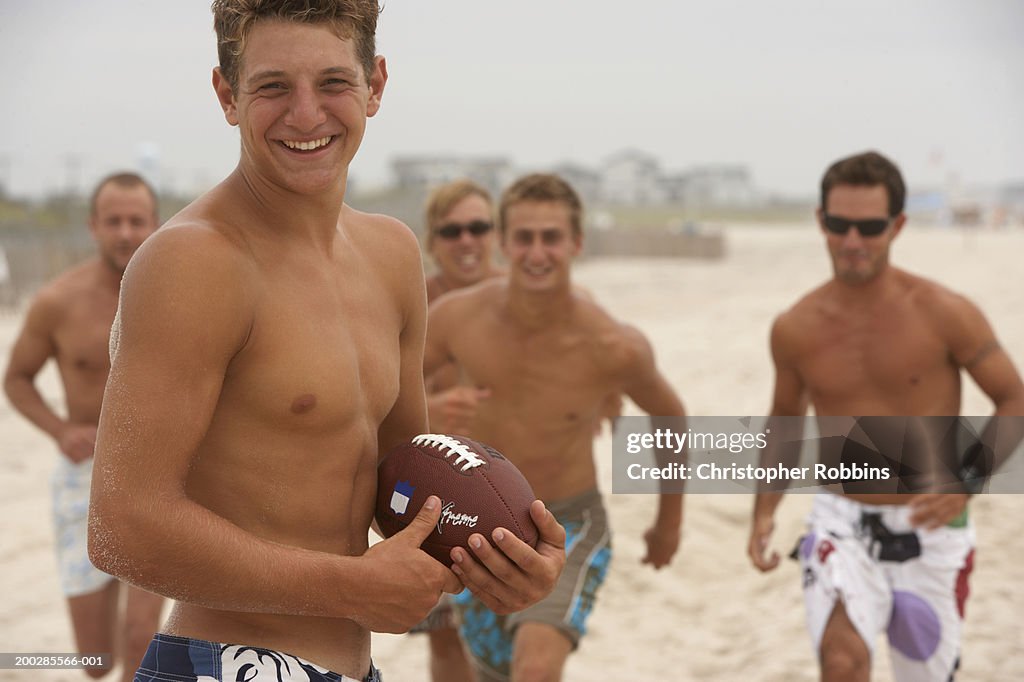Young man holding football on beach four men running in background
