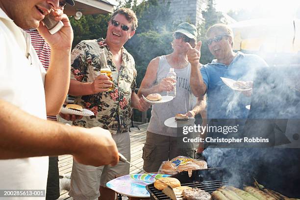man barbecuing using mobile phone, friends laughing in background - papieren bord stockfoto's en -beelden