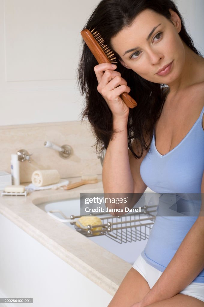 Young woman perched on edge of bath, brushing hair, close-up