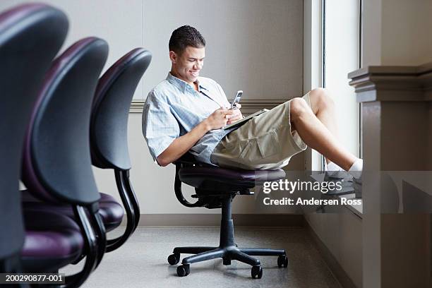 young man sitting on office chair, using cell phone, side view - korte broek stockfoto's en -beelden
