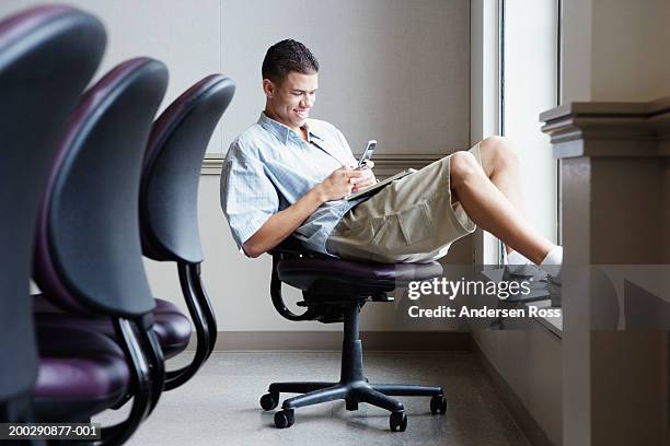 young man sitting on office chair, using cell phone, side view - shorts stockfoto's en -beelden