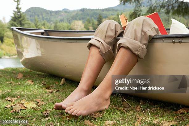 barefoot boy (9-11 years) reading book in canoe by lake, low section, close-up of legs over edge of canoe - rolled up trousers stock-fotos und bilder