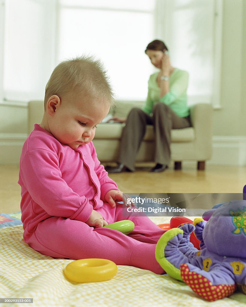 Baby girl (6-9 months) playing with toys, mother in background