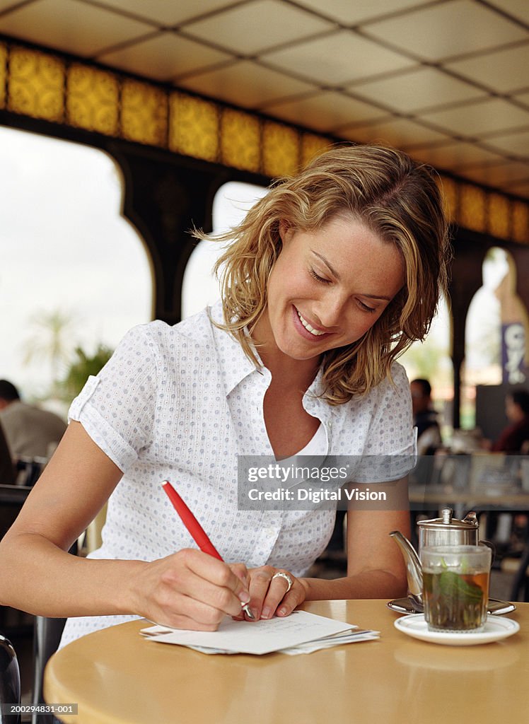 Young woman at table writing on envelope by teapot and glass, smiling