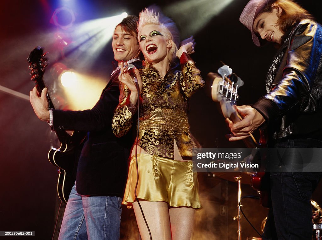 Woman holding microphone flanked by men playing guitars, smiling