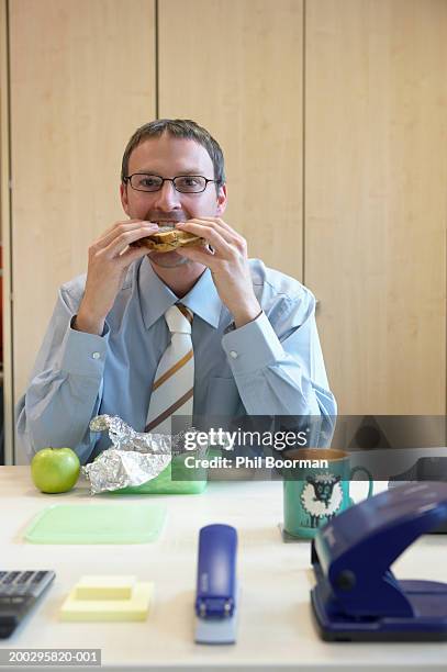 young businessman eating sandwich at desk, portrait - staples office stockfoto's en -beelden