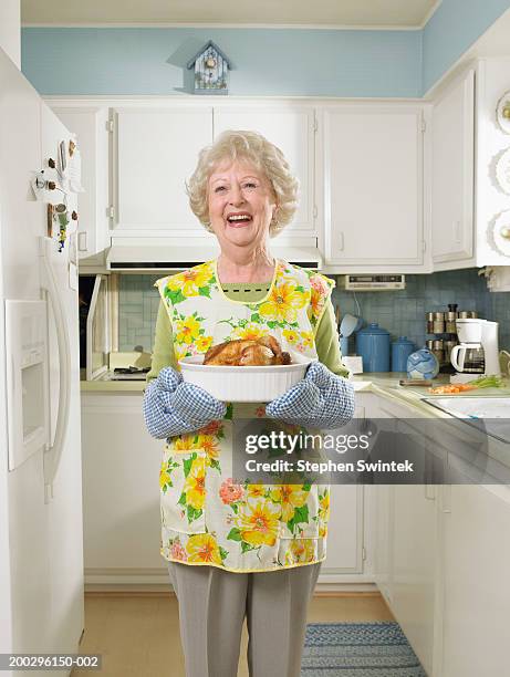 senior woman in kitchen,  holding dish with roasted chicken - kitchen apron stock pictures, royalty-free photos & images