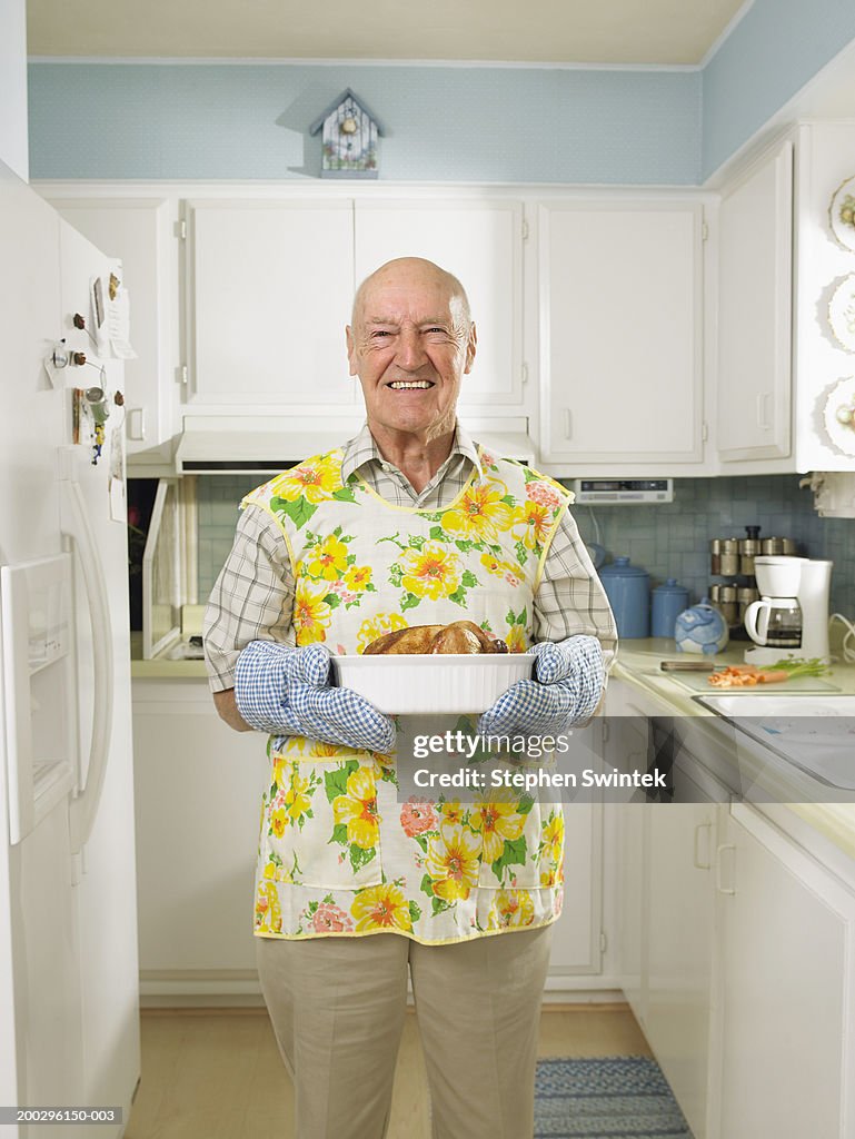 Senior man in kitchen,  holding dish with roasted chicken