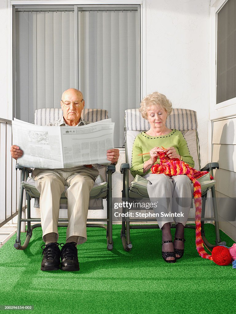 Senior couple sitting on patio, man reading paper, woman knitting