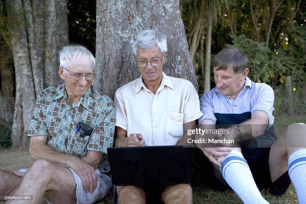 Mature man and senior men by tree, looking at laptop screen, smiling
