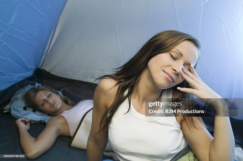 Young woman waking up in tent, eyes closed, woman in background