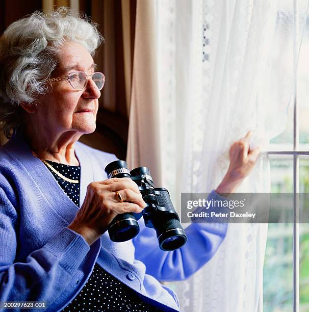 senior woman looking out window, holding binoculars - spying neighbor photos et images de collection