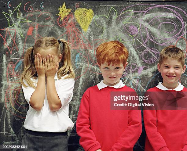 three children (6-8) standing by blackboard, girl covering face - redhead boy fotografías e imágenes de stock