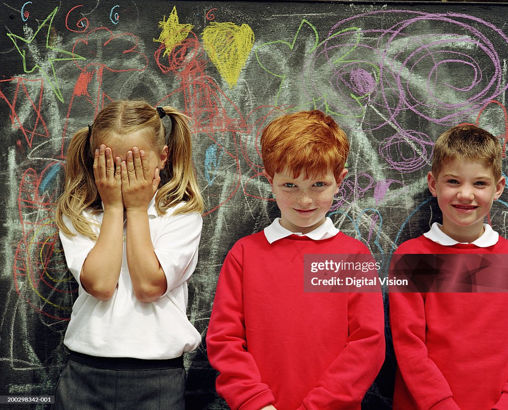 Three children (6-8) standing by blackboard, girl covering face