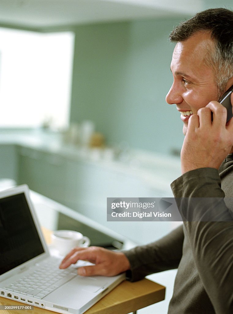 Mature man in kitchen with laptop, using mobile phone, profile