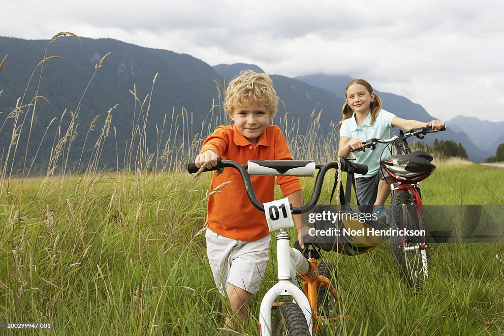 Boy and girl (5-8 years) with mountain bikes in long grass, smiling, portrait