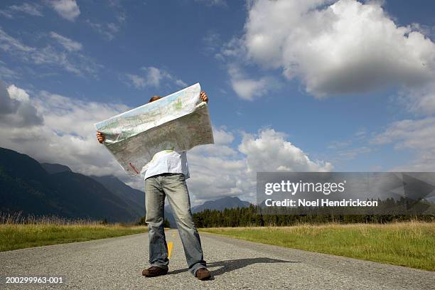 man standing on rural road holding road map, head obscured by map - roadmap stock-fotos und bilder