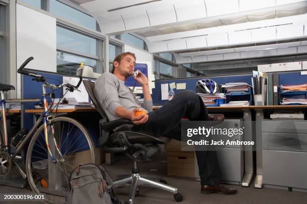 young man relaxing at office desk, using telephone and holding orange - gambe accavallate foto e immagini stock