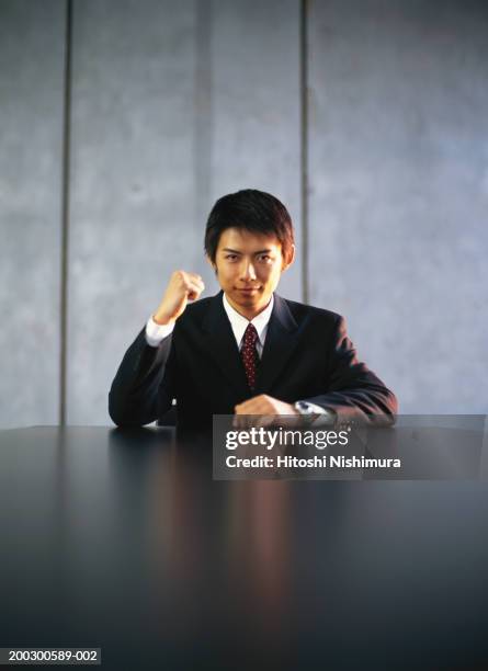 young businessman holding up fist at office table, portrait - faust in die luft stoßen stock-fotos und bilder