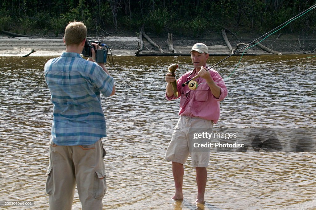 Man filming man holding fishing rod and fish on river