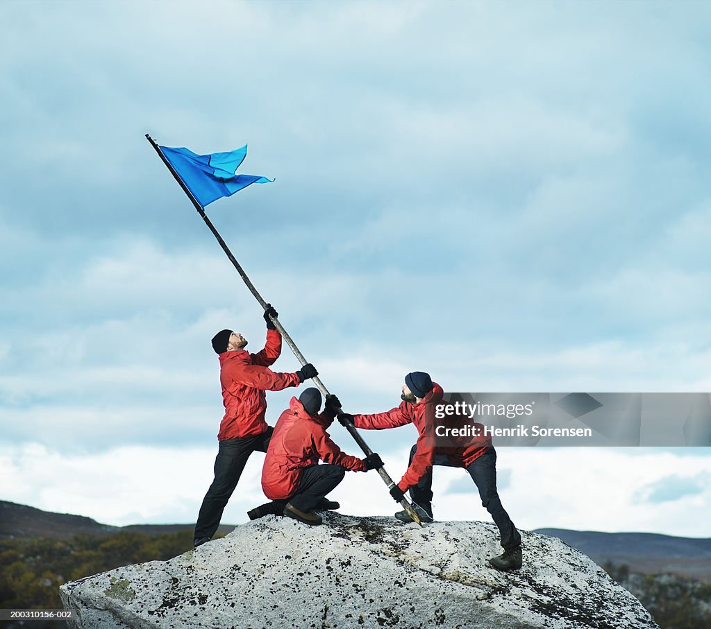 Three male hikers raising flag on rock
