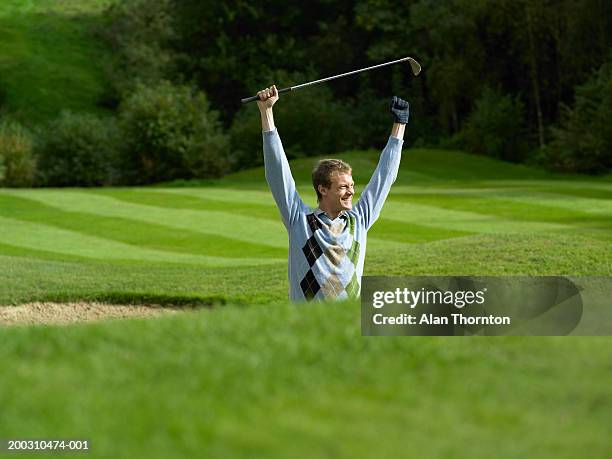 male golfer standing in bunker holding golf club, arms raised, smiling - golf club stock-fotos und bilder