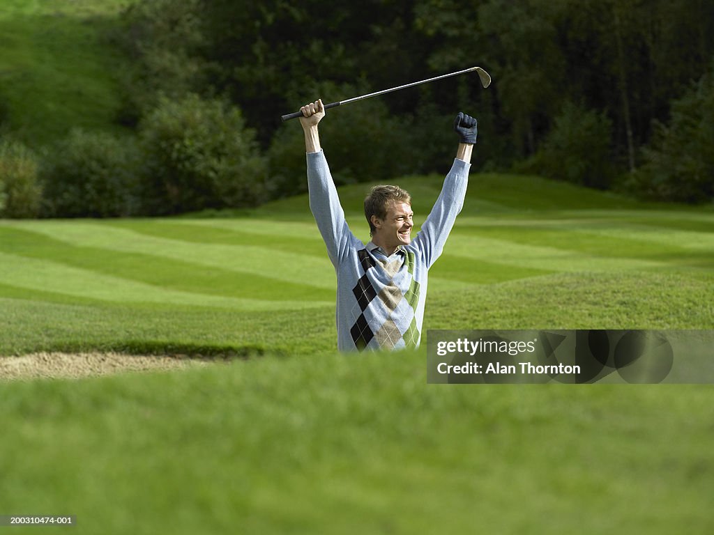Male golfer standing in bunker holding golf club, arms raised, smiling