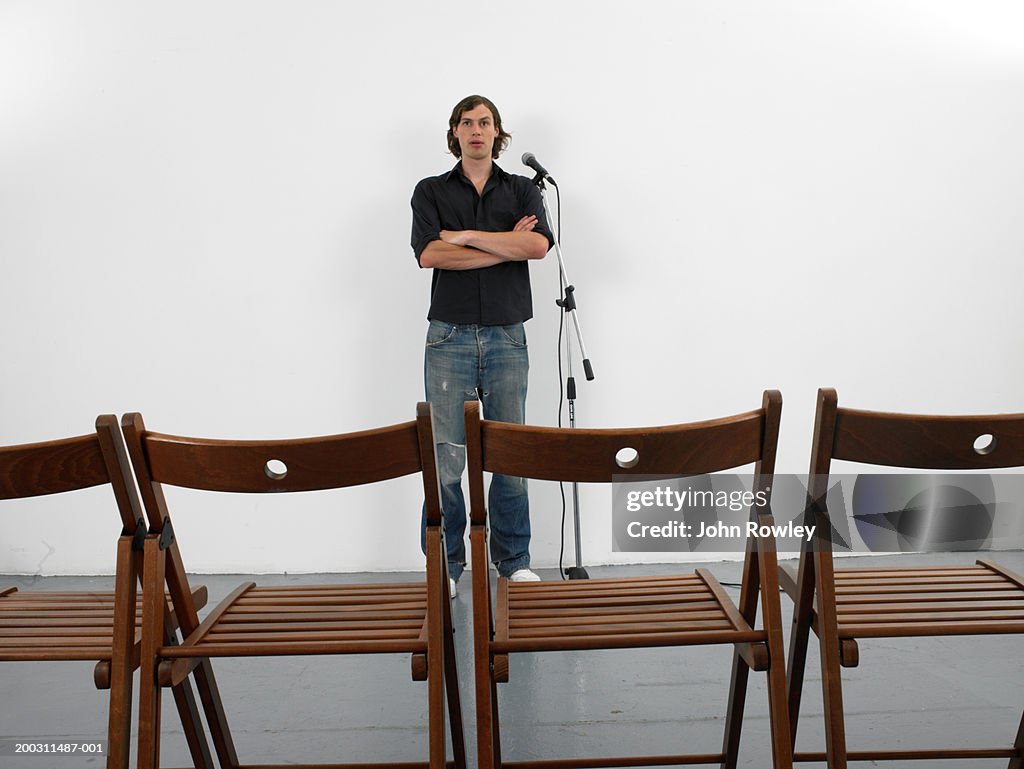 Man standing by microphone facing row of empty chairs, arms crossed