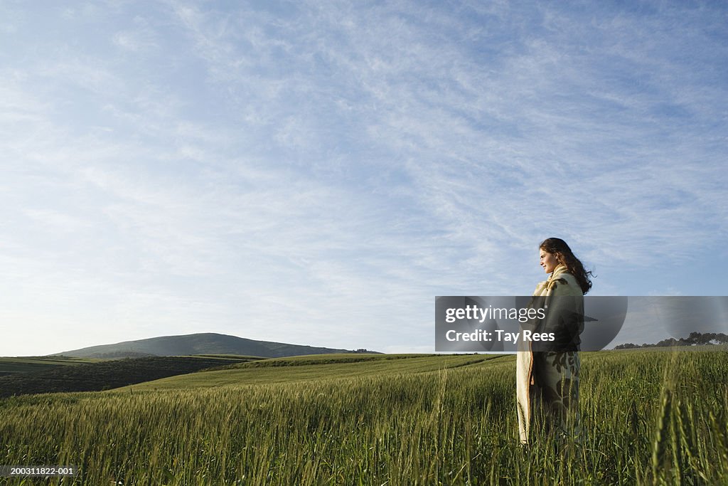 Woman standing in field, wrapped in blanket, side view