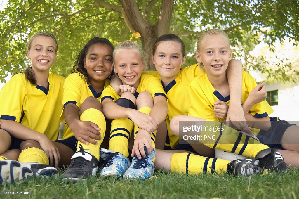 Five girls (10-17) in soccer uniforms sitting on field, portrait