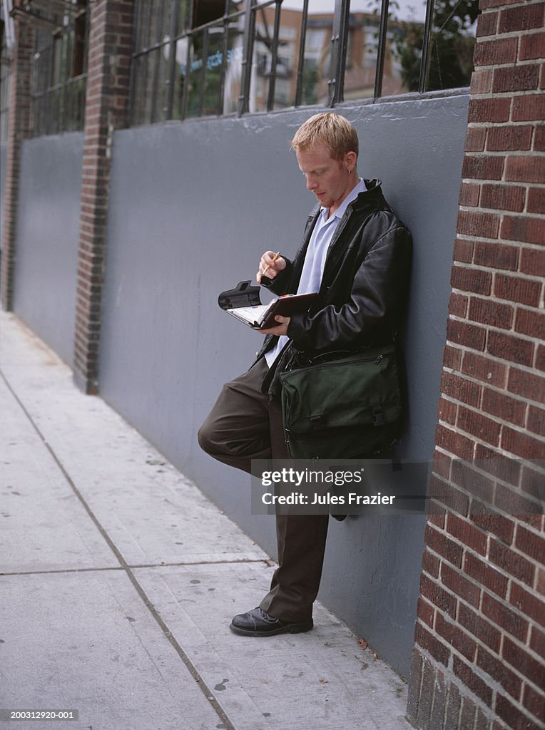 Man leaning against wall outdoors, holding file binder