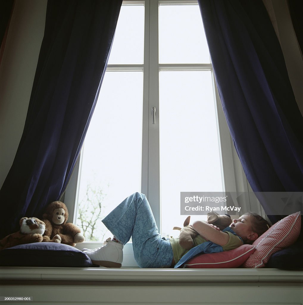 Girl (6-7) lying on window sill, hugging toy dog