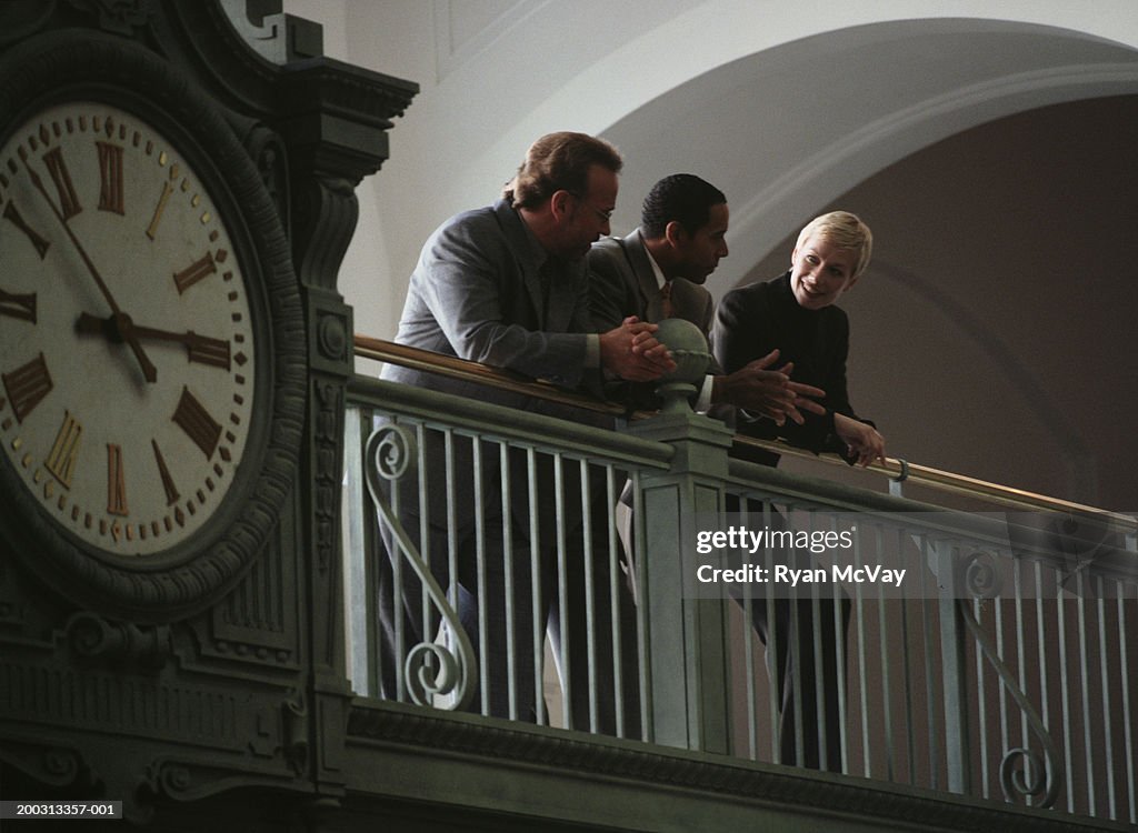 Three adult leaning over interior railing, large clock in foreground low angle view