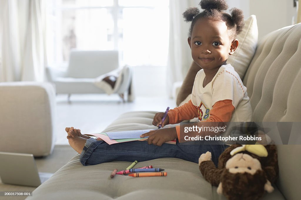 Girl (2-4) on sofa, holding crayon and paper, smiling, portrait