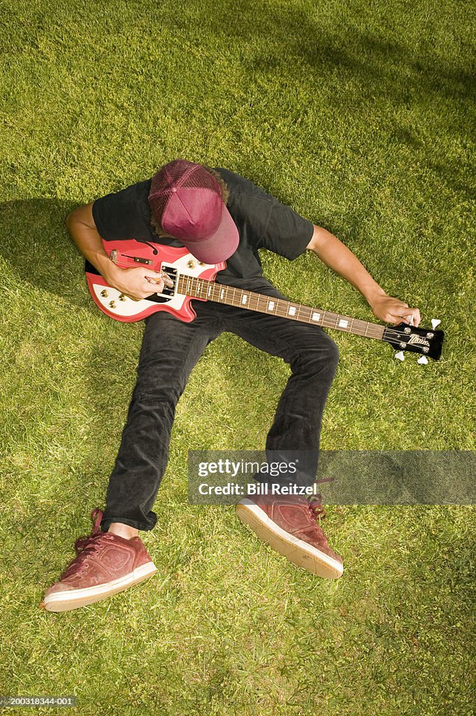 Teenage boy (13-15) sitting on lawn, tuning bass guitar, elevated view