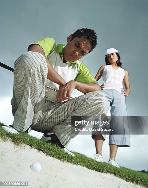 young man frowning at golf ball in sand trap, young woman looking on - golf bunker low angle stock pictures, royalty-free photos & images