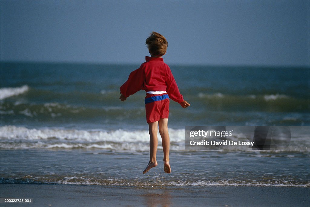 Boy (4-7) jumping in water's edge, rear view